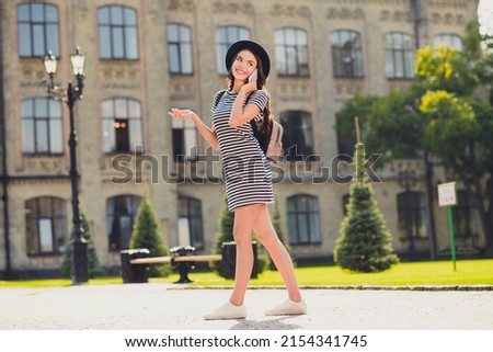 Photo of cute adorable young woman dressed striped outfit cap backpack walking talking modern device smiling outside landscape