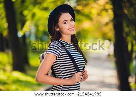 Photo of charming pretty young lady wear striped dress headwear rucksack smiling walking enjoying good weather outdoors urban city park