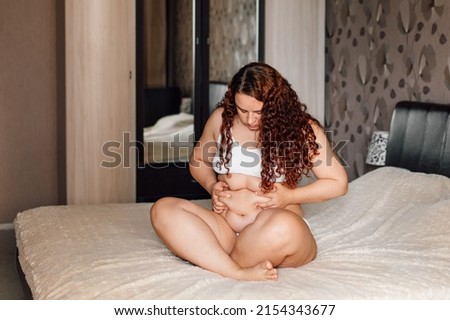 Curly plus size woman in white underwear sitting on bed in bedroom and pinch sagging folds on thick stomach. Fat burning treatment of overweight body. Weight loss program and anticellulite massage.