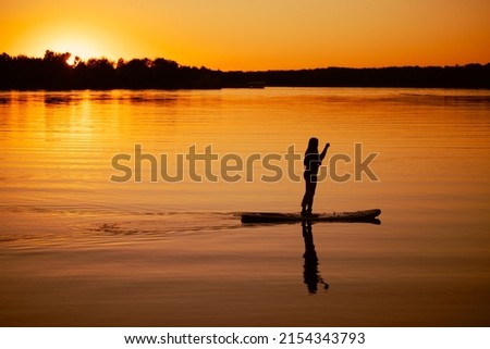 Female paddle boarder moving around with oar in hands on lake with dazzling sunset in background and reflection on surface water in summer. Active lifestyle.