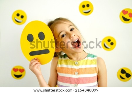 The girl holds a sad emoticon  face in hand, but she smiling and laughs no matter what. A cheerful happy sincere child