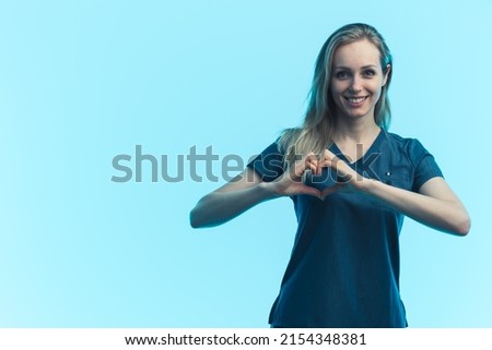 Young adult attractive joyful female nurse wearing a blue medical uniform showing a heart gesture with her hands. Studio shot. High quality photo