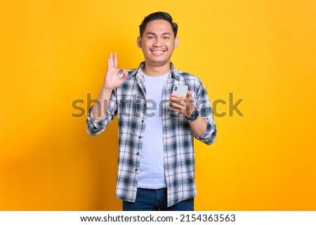 Cheerful young Asian man in plaid shirt using mobile phone and showing okay sign gesture, recommend application isolated on yellow background