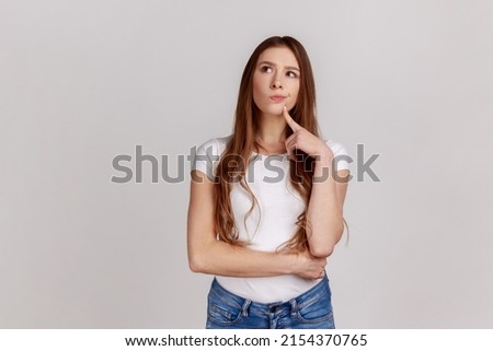 Portrait of standing with thoughtful serious smart expression, pondering answer, having doubts and suspicion, wearing white T-shirt. Indoor studio shot isolated on gray background. Royalty-Free Stock Photo #2154370765