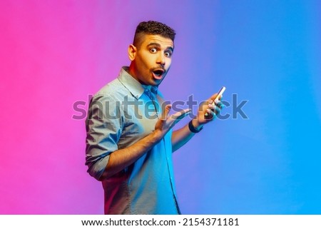Side view of astonished surprised man in shirt using online service on mobile phone and expressing amazement, looking at camera. Indoor studio shot isolated on colorful neon light background.