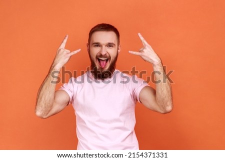 Portrait of bearded man showing rock and roll sign hand gesture, looking at camera with tongue out, excited expression, wearing pink T-shirt. Indoor studio shot isolated on orange background.