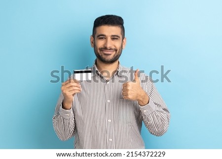 Businessman holding credit card, looking with toothy smile, recommend bank or shopping discounts, showing thumb up, wearing striped shirt. Indoor studio shot isolated on blue background. Royalty-Free Stock Photo #2154372229