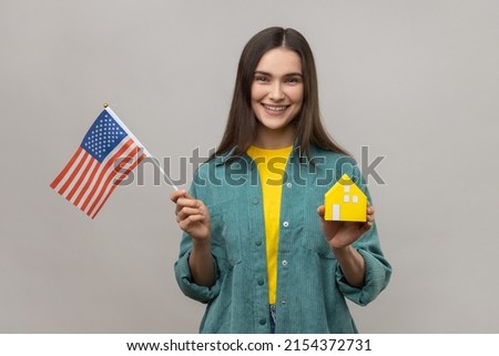 Portrait of delighted smiling holding american flag and paper house, dreaming to buy accommodation in USA, wearing casual style jacket. Indoor studio shot isolated on gray background.