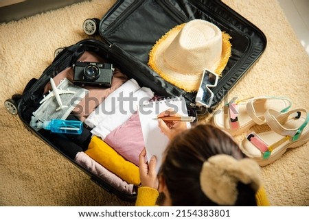 Making check list of things to pack for travel. Woman writing paper take note and packing suitcase to vacation writing paper list sitting on room, prepare clothes into luggage, Travel vacation travel Royalty-Free Stock Photo #2154383801