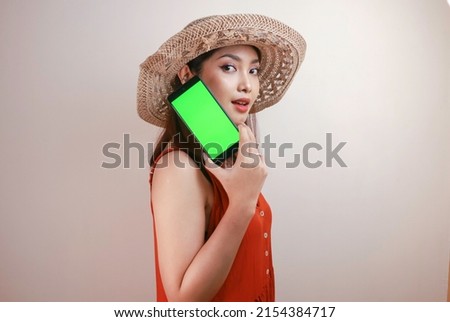 Excited and smiling young Asian woman pointing finger at smartphone in her hand, emotionally reacting to new app, overjoyed millennial woman standing isolated over white studio background