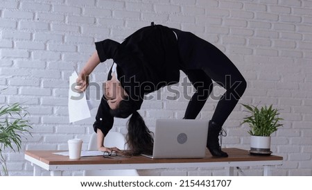 An employee of the office right on the desktop performed a complex exercise - stood on the bridge. At this point, she's inspired to look through the documents. She is dressed in stylish black clothes.