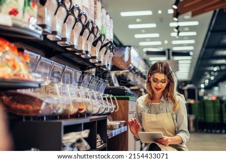 Young woman, employee with apron holding digital tablet and checking condition of product in the store
