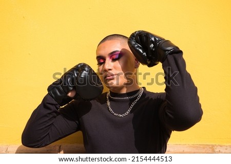Young non-binary person wears boxing gloves on a mustard yellow background, the person is make up and does different boxing poses. Concept equality, homosexuality, gay, lesbian, gay pride. Royalty-Free Stock Photo #2154444513
