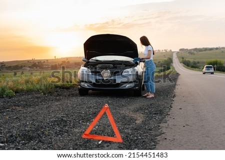 A young girl stands near a broken car in the middle of the highway during sunset and tries to repair it. Troubleshooting the problem. Waiting for help. Car service. Car breakdown on road.
 Royalty-Free Stock Photo #2154451483
