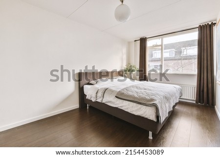 Comfortable bed and lamp placed in small narrow minimalist style bedroom with white walls and brown curtains on window above radiator in modern apartment Royalty-Free Stock Photo #2154453089