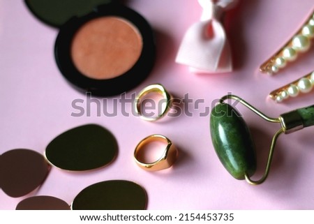 Pink scrunchie, gold earrings and rings, blush, various hair barrettes, lipstick and jade roller on bright pink background. Selective focus.