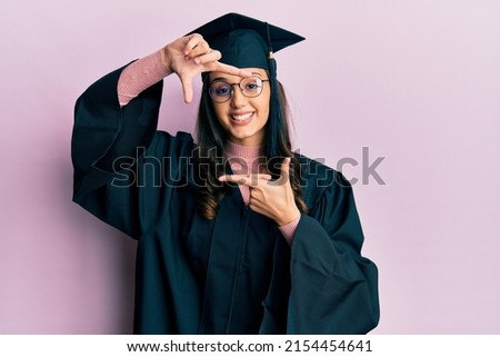 Young hispanic woman wearing graduation cap and ceremony robe smiling making frame with hands and fingers with happy face. creativity and photography concept. 