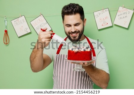 Young fun male chef confectioner baker man wear striped apron hold adorn birthday sweet dessert cake with cream isolated on plain pastel light green background studio portrait. Cooking food concept. Royalty-Free Stock Photo #2154456219
