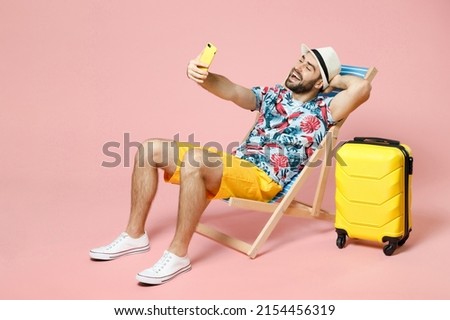 Full length cheerful young traveler tourist man in hat sit on deck chair doing selfie shot on mobile phone isolated on pink background studio. Passenger travel on weekend. Air flight journey concept