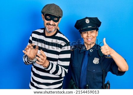 Middle age couple of hispanic woman and man wearing thief and police uniform doing happy thumbs up gesture with hand. approving expression looking at the camera showing success. 