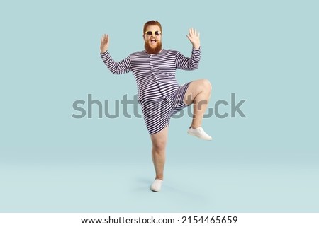 Funny fat guy enjoying his happy summer holiday. Full body length portrait of cheerful goofy chubby bearded man wearing striped retro swimsuit and sunglasses dancing against blue studio background Royalty-Free Stock Photo #2154465659