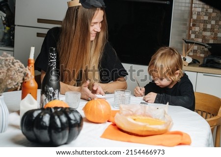 Mom and her little son sitting behind a round table, preparing decorations for halloween. Painting on stones, pumpkins and bottles.
