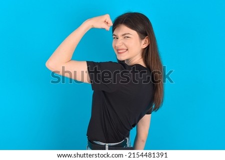 Young caucasian woman wearing black T-shirt over blue background,  showing muscles after workout. Health and strength concept. Royalty-Free Stock Photo #2154481391