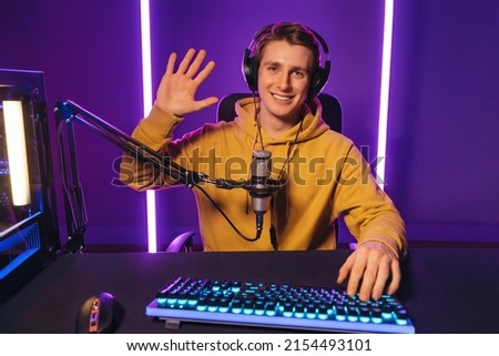 Young handsome vlogger pro gamer waving hand to camera, says hello to his subscribers and followers, smiling on camera. He recording live stream of video game playthrough at home at night. Cyber sport Royalty-Free Stock Photo #2154493101