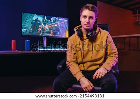 Portrait of young serious pro gamer wearing headset sitting at desk with gaming equipment, looking at camera. Neon coloured room background. Cyber sport, e-Sport and video games addiction concept