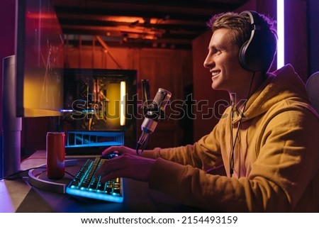  Excited caucasian gamer streamer with headphones playing computer pc video at his living room, recording live stream using microphone, drinking energy drink. Cyber sport and social media concept