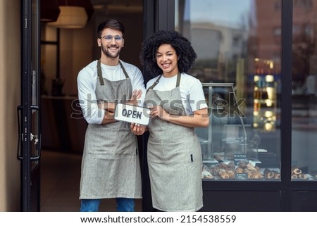 Portrait of two owners friends holding a open sign together while smiling to the camera in front of pastry shop.