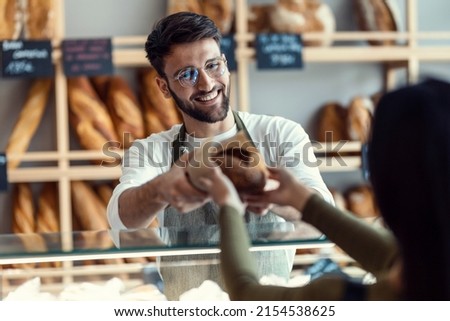 Shot of cheerful seller giving fresh loaf of bread to smiling woman in the pastry shop. Royalty-Free Stock Photo #2154538625