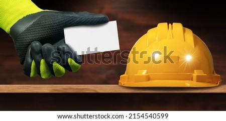 Closeup of a yellow Safety Helmet above a wooden workbench and a Manual worker with green and black protective work gloves holding a blank business card with copy space.
