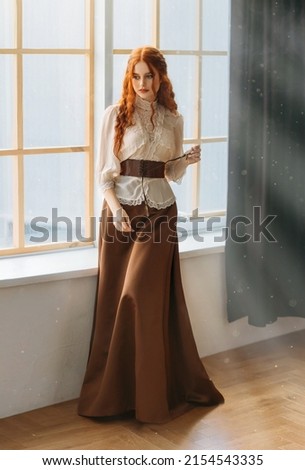 Red-haired woman in vintage dress stands at large classic window waiting love. Clothing costume countess old style white blouse, brown long skirt. Curly red hair. Redhead girl princess 1800s stylish. Royalty-Free Stock Photo #2154543335