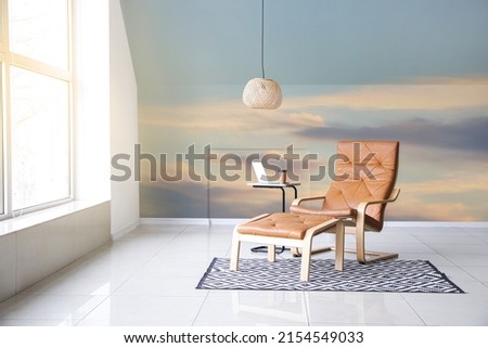 Comfortable armchair with table and chandelier near wall with printed sky in room