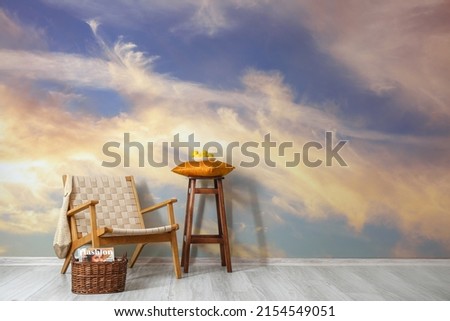 Stylish wicker armchair with basket and stool near wall with printed sky in room