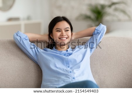 Cute young chinese woman in blue shirt and jeans resting on sofa at home, leaning back, looking at copy space and smiling, dreaming about vacation, cozy living room interior