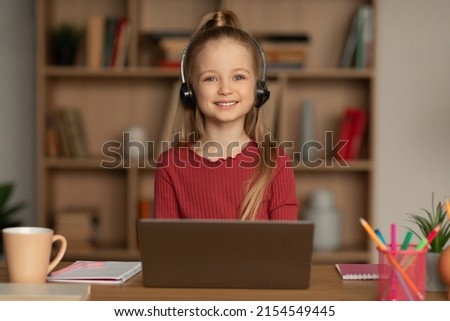 Cheerful School Girl Using Laptop Wearing Earphones Smiling To Camera Learning Online At Home. Educational Website, Internet And Remote Education. Front View, Selective Focus