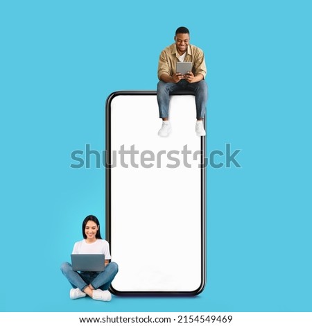 Great App. Excited Black Guy And Lady Sitting On Big Giant Smartphone With Blank White Screen Using Gadgets, Cheerful Couple Chatting On Social Media, Blue Wall, Mock Up. Modern Communication Concept Royalty-Free Stock Photo #2154549469