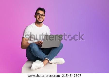 Young Arab man in casual wear working online, sitting cross legged and using laptop in neon light, full length. Millennial Eastern guy surfing internet on portable pc, copy space