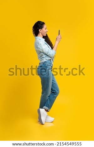 Profile portrait of happy smiling young woman using cell phone standing isolated on yellow orange wall. Female chatting online, browsing social media, taking photo, full body length, side view