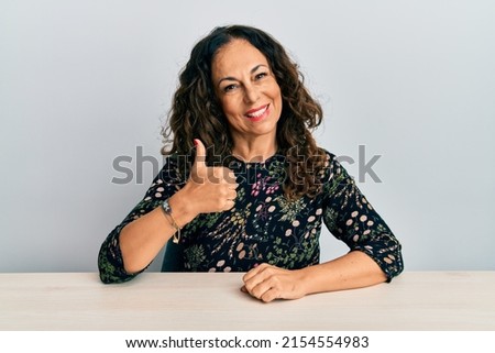 Beautiful middle age woman wearing casual clothes sitting on the table doing happy thumbs up gesture with hand. approving expression looking at the camera showing success. 