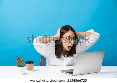 Surprised hispanic woman looking at laptop hands on head while sitting behind desk isolated on blue background. Royalty-Free Stock Photo #2154566725