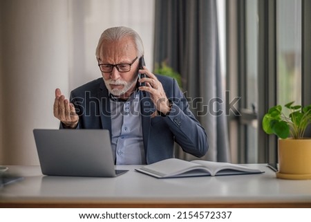 Anxious businessman talking on mobile phone while working on laptop, Worried senior male professional is wearing formals, He is sitting at desk in office.