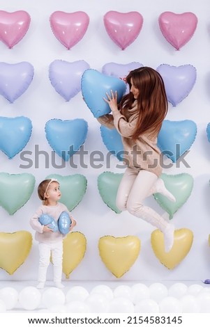 Beautiful young mother and little cute daughter having fun together stand on colorful heart balloons background and holding blue Valentine's balloon. birthday, velentines, mothers day.