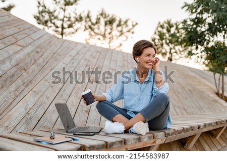 young pretty woman working on laptop in IT outside in modern park street sitting on remote work, summer style casual smiling talking on wireless earpods drinking coffee Royalty-Free Stock Photo #2154583987