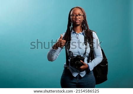 Joyful photographer giving ok finger gesture while standing on blue background. Cheerful professional photography entusiast with DSLR device saying is ready for photo shooting by giving thumbs up sign