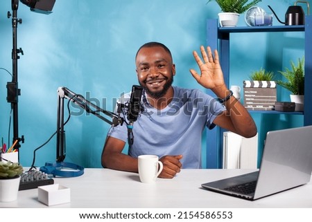 Smiling vlogger waving hello sitting at desk in recording studio with professional microphone and video light. Content creator doing hand gesture in front of audio podcast setup.