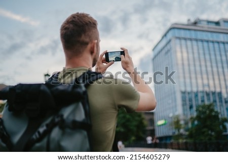 handsome hipster man walking in street with urban backpack traveler in khaki colors, using smartphone camera taking photo