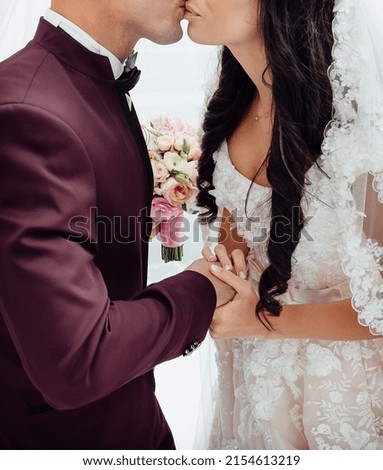 The bride and groom kiss at the wedding ceremony. The groom is in a crimson suit, the bride is dressed in a light dress with patterns, with a bouquet in her hands.